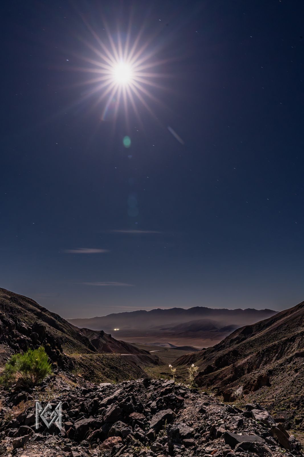 A starburst moon over a rocky canyon, the wash and distant valley glowing below in moonlight
