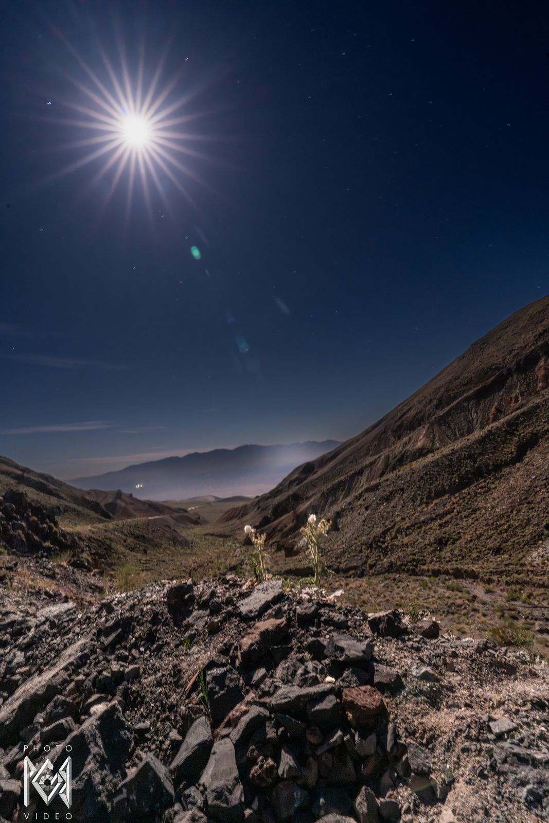 Looking up a boulder-strewn slope at a starburst moon, with a canyon and hazy valley beyond