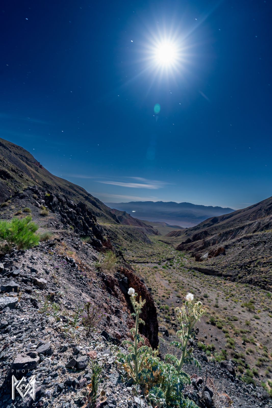 A blazing moon in a deep blue sky above a rocky canyon and a green-flecked slope, valley and mountains beyond