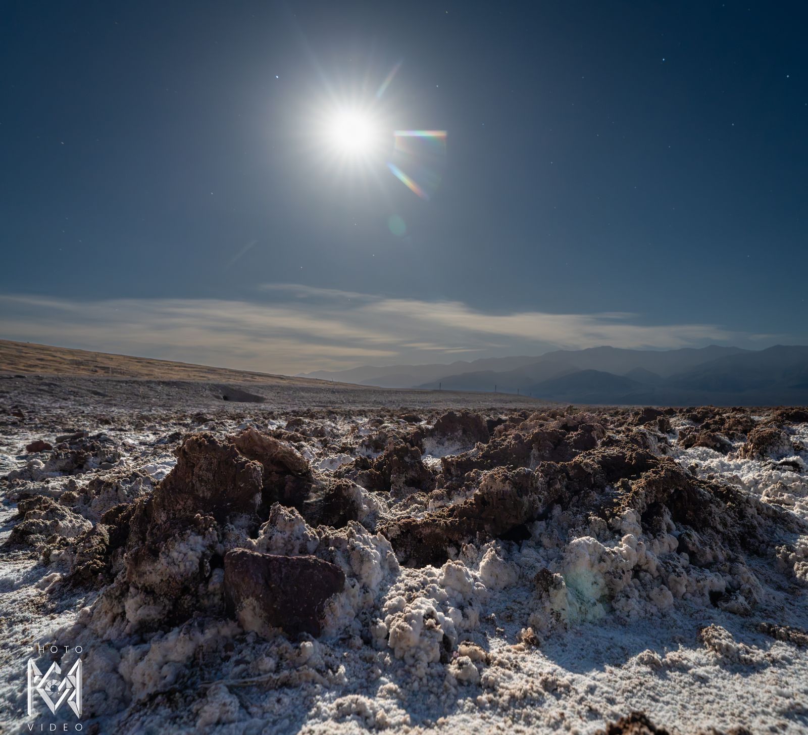 Moonlight on crusted white salt and mud mounds, with the moon above a layer of cloud over the distant range
