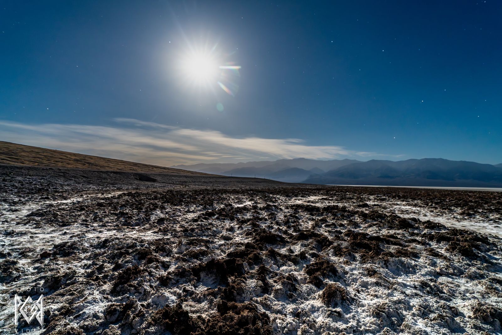 The moon above cloud over a salt flat of crumbling white crust, mountains on the horizon