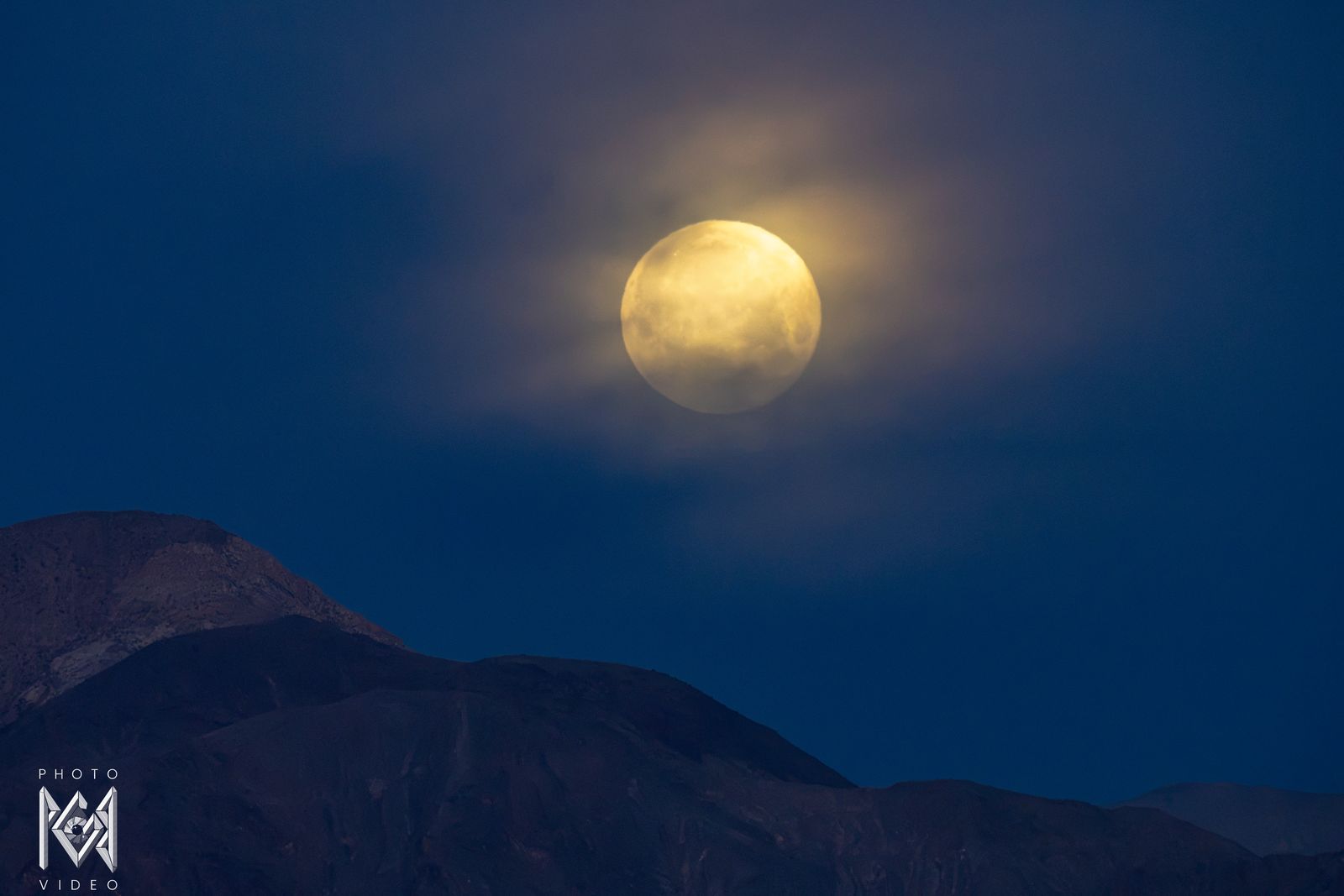 A golden moon wrapped in cloud in a deep blue sky above dark mountain ridges