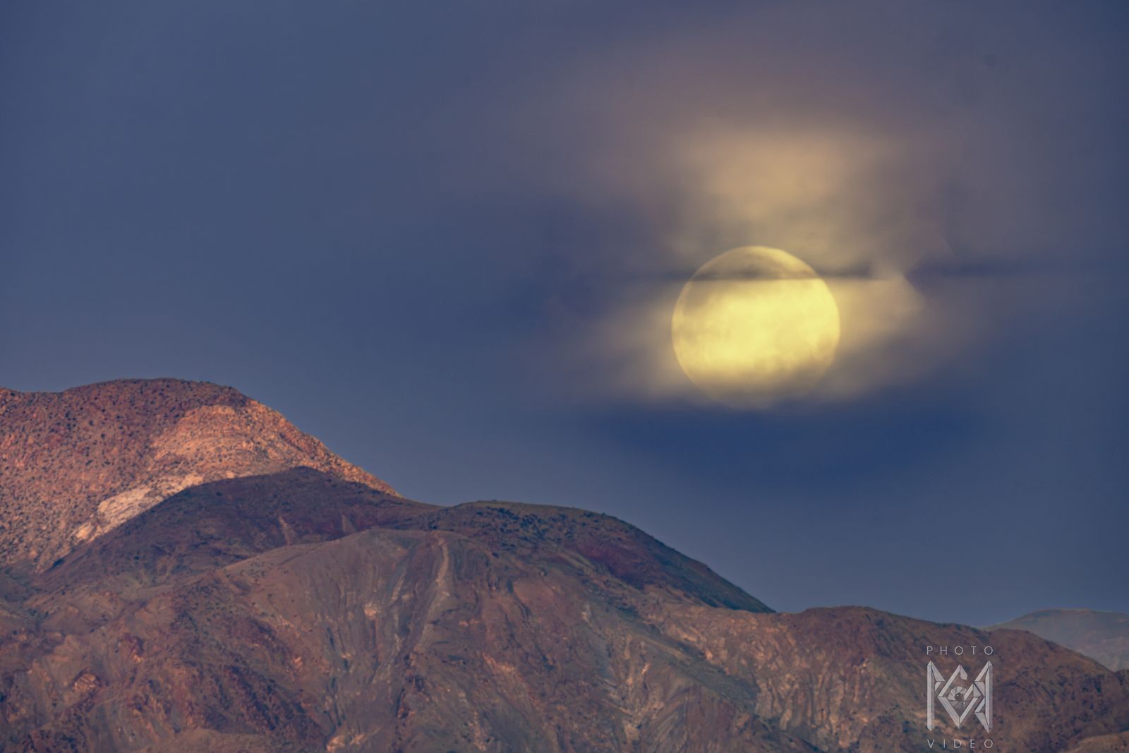 A pale yellow moon in glowing cloud above rust-red mountain ridges