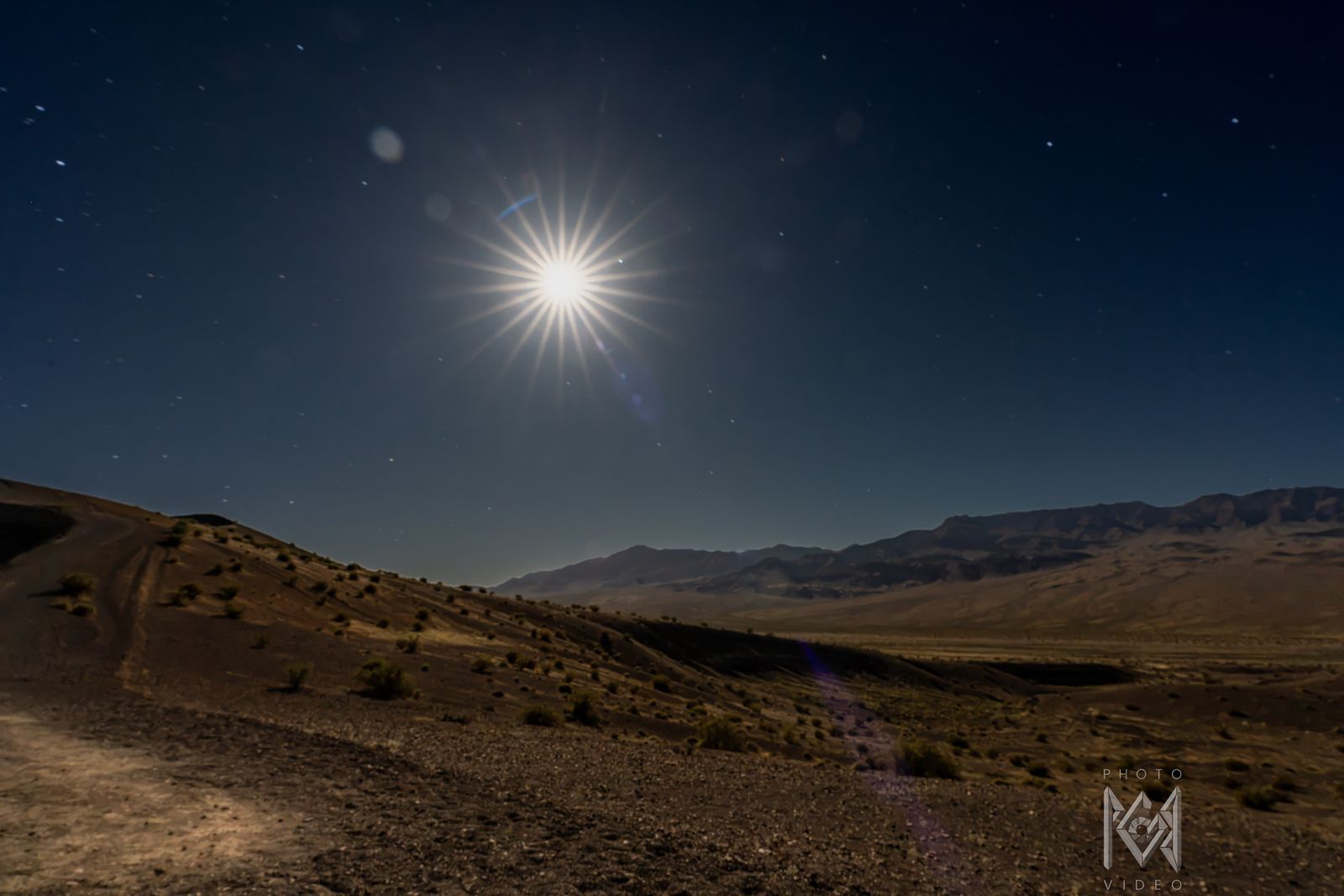 A starburst moon above a dirt track curving into a wide desert valley and distant ranges