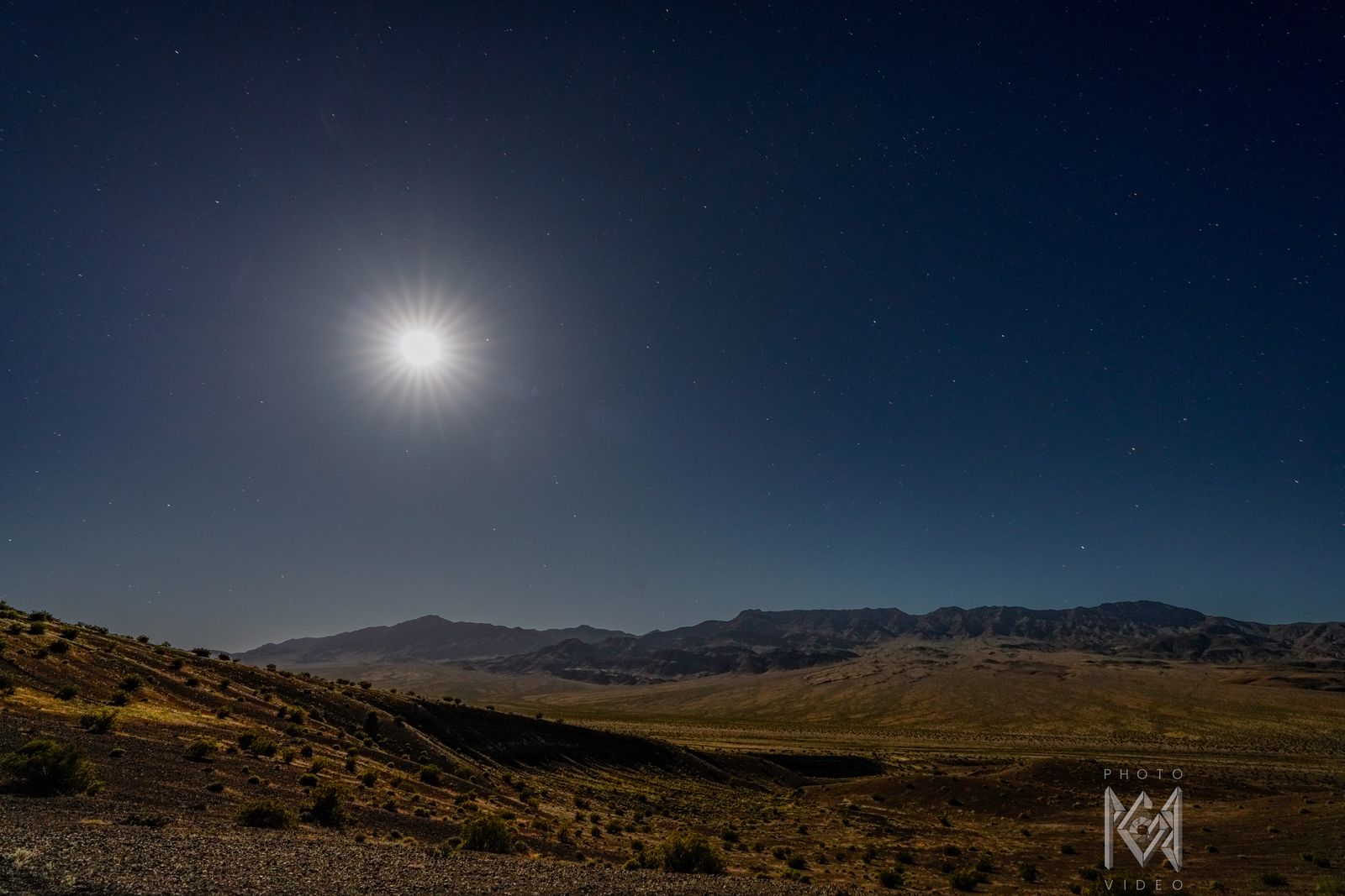 A bright moon in a starry sky above a dark valley of shrubs and the long ridge beyond