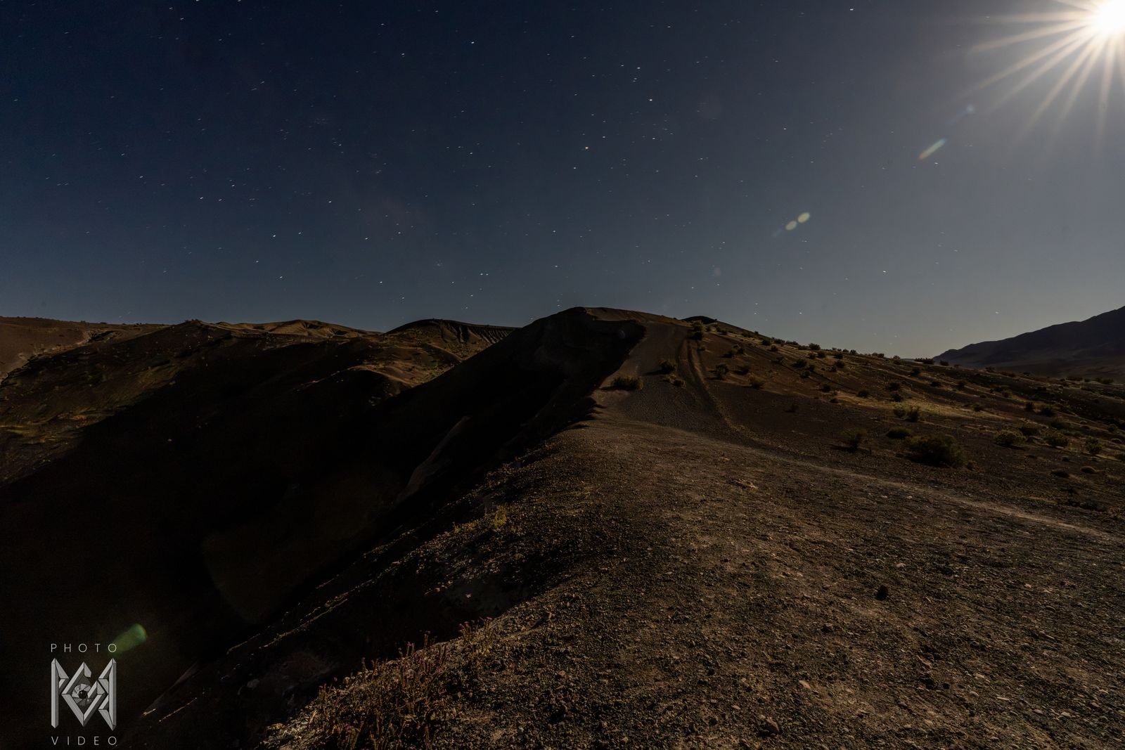 A dark ridge with a track along its crest under a starry sky, the moon's flare in the top corner