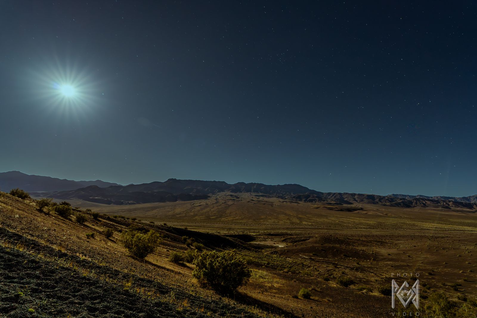 A pale blue starburst moon over a moonlit valley of golden shrubs and distant ranges