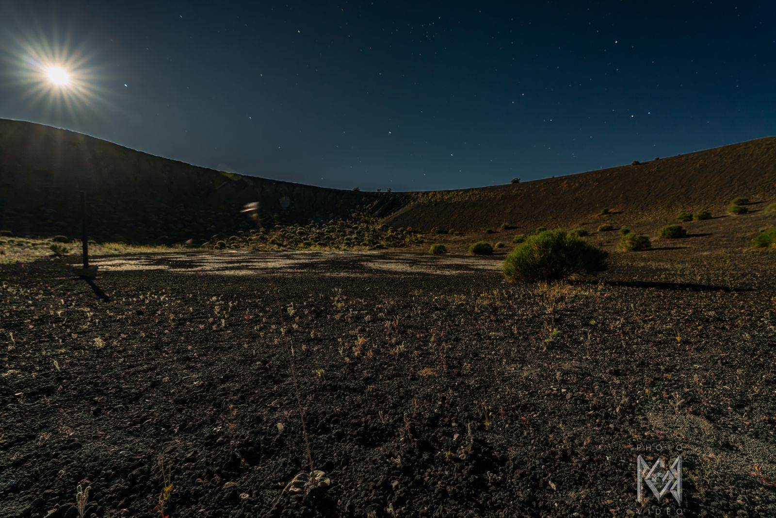 The moon flaring at the top left above a dark hillside and a pale gravel flat with a shrub, under a starry sky