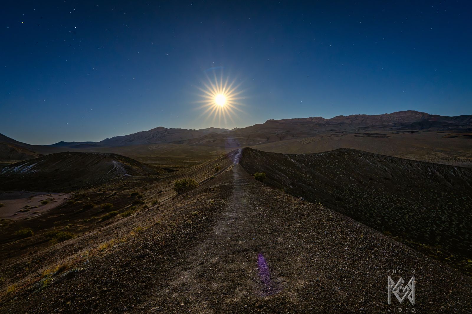 The moon low over a long ridge and a path leading to the horizon, golden light on the hills and a blue dusk sky
