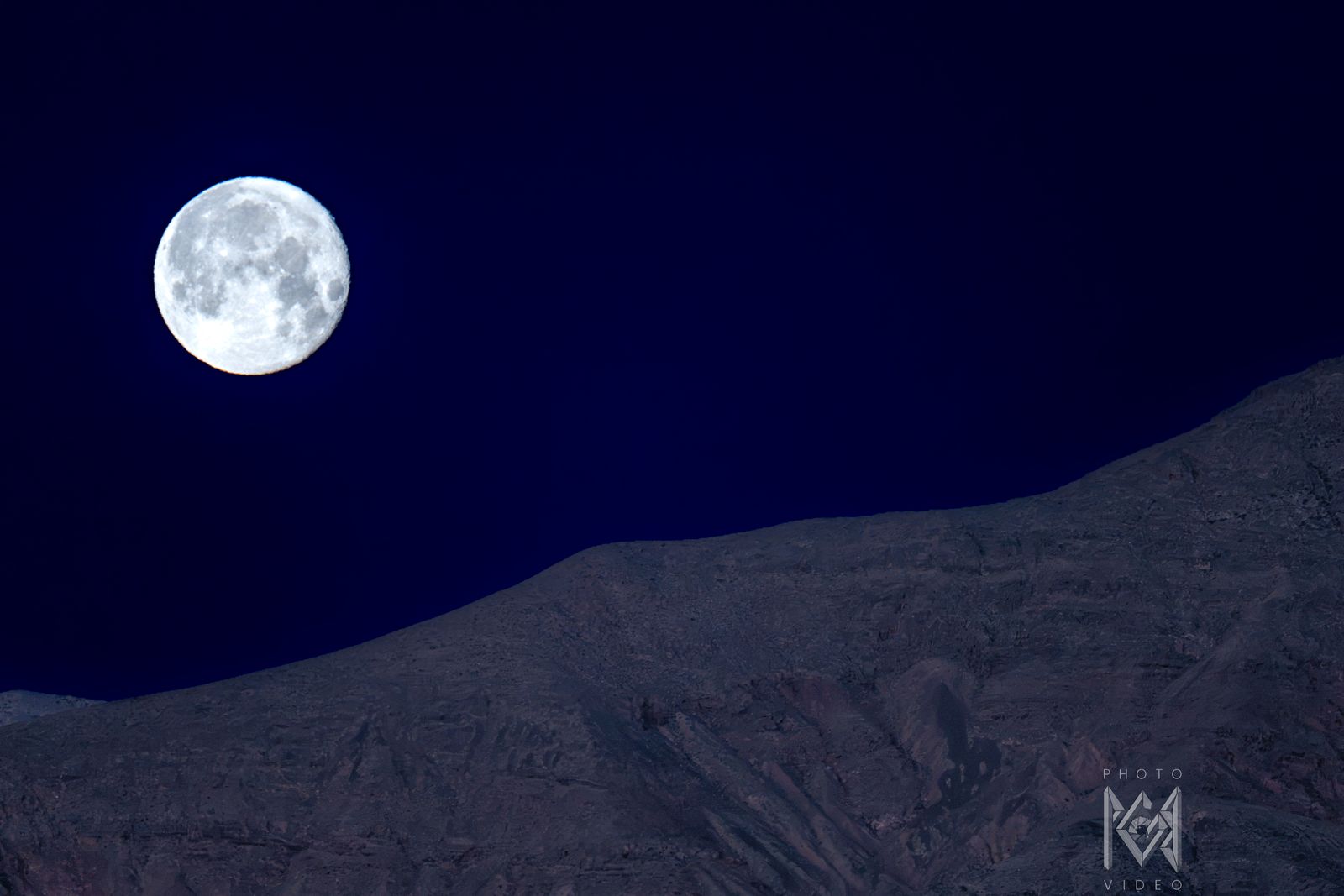 A bright full moon in a deep navy sky above a dim, softly lit ridge