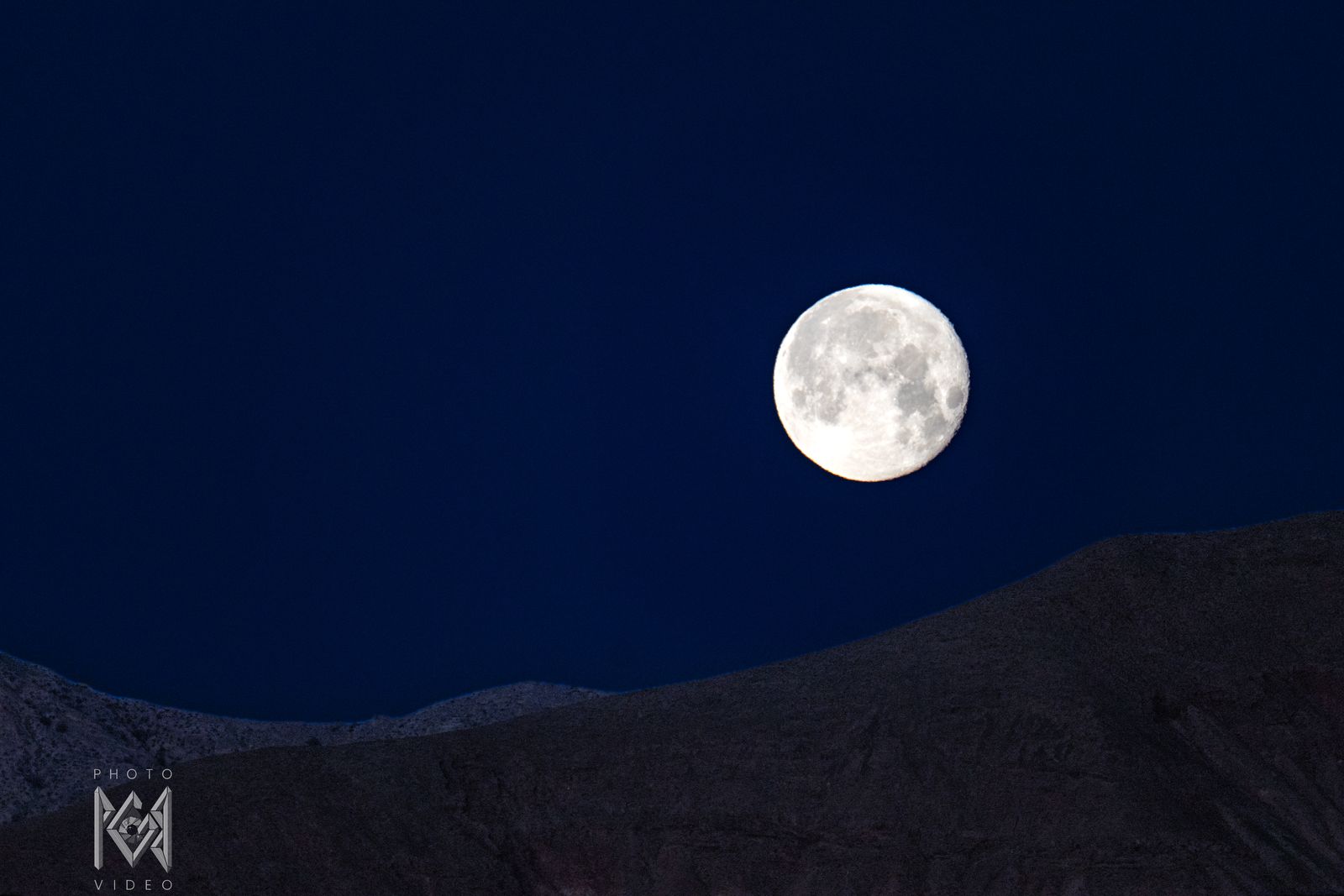 A bright full moon in a deep navy sky above a dark, softly lit ridge