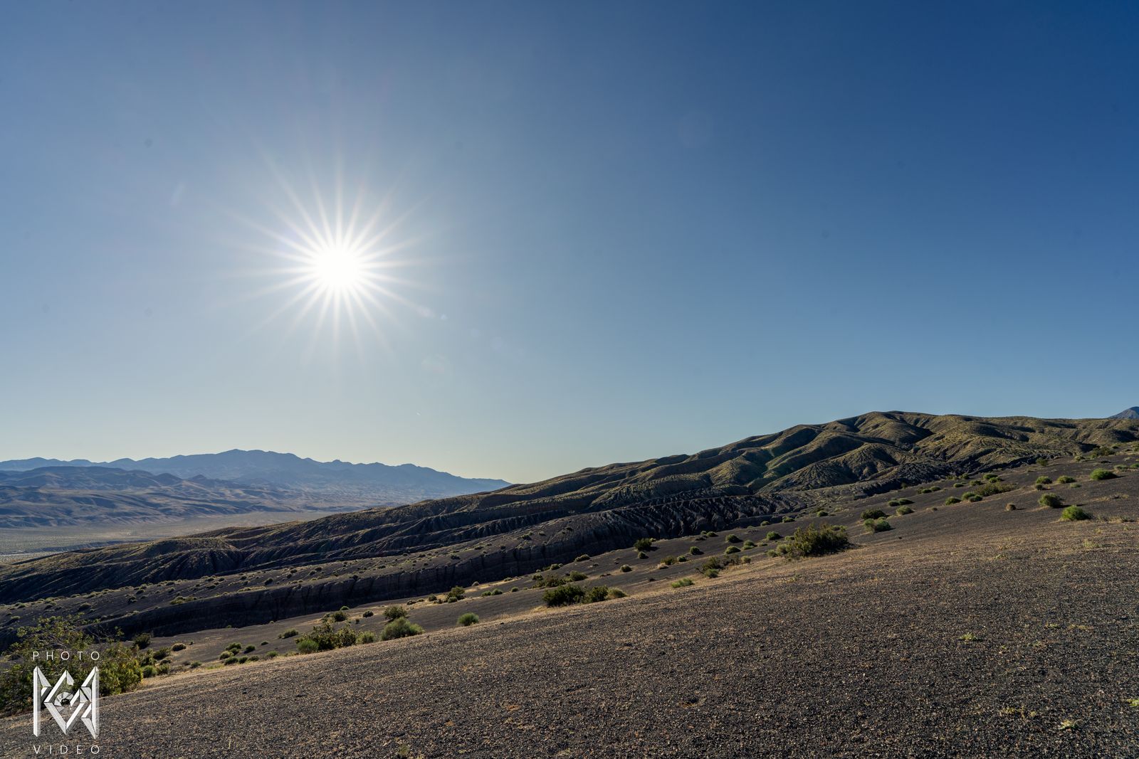 A starburst moon in a pale blue morning sky above a dune-like ridge and a gravel plain, mountains behind