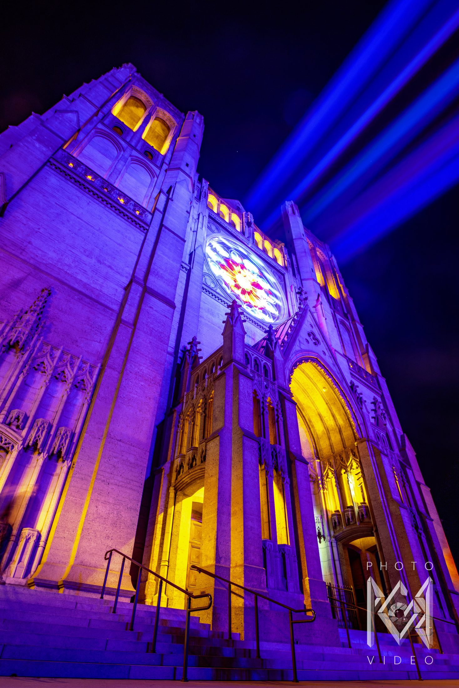 Looking up at a Gothic cathedral facade lit violet and gold, with a glowing rose window and blue beams of light crossing the night sky