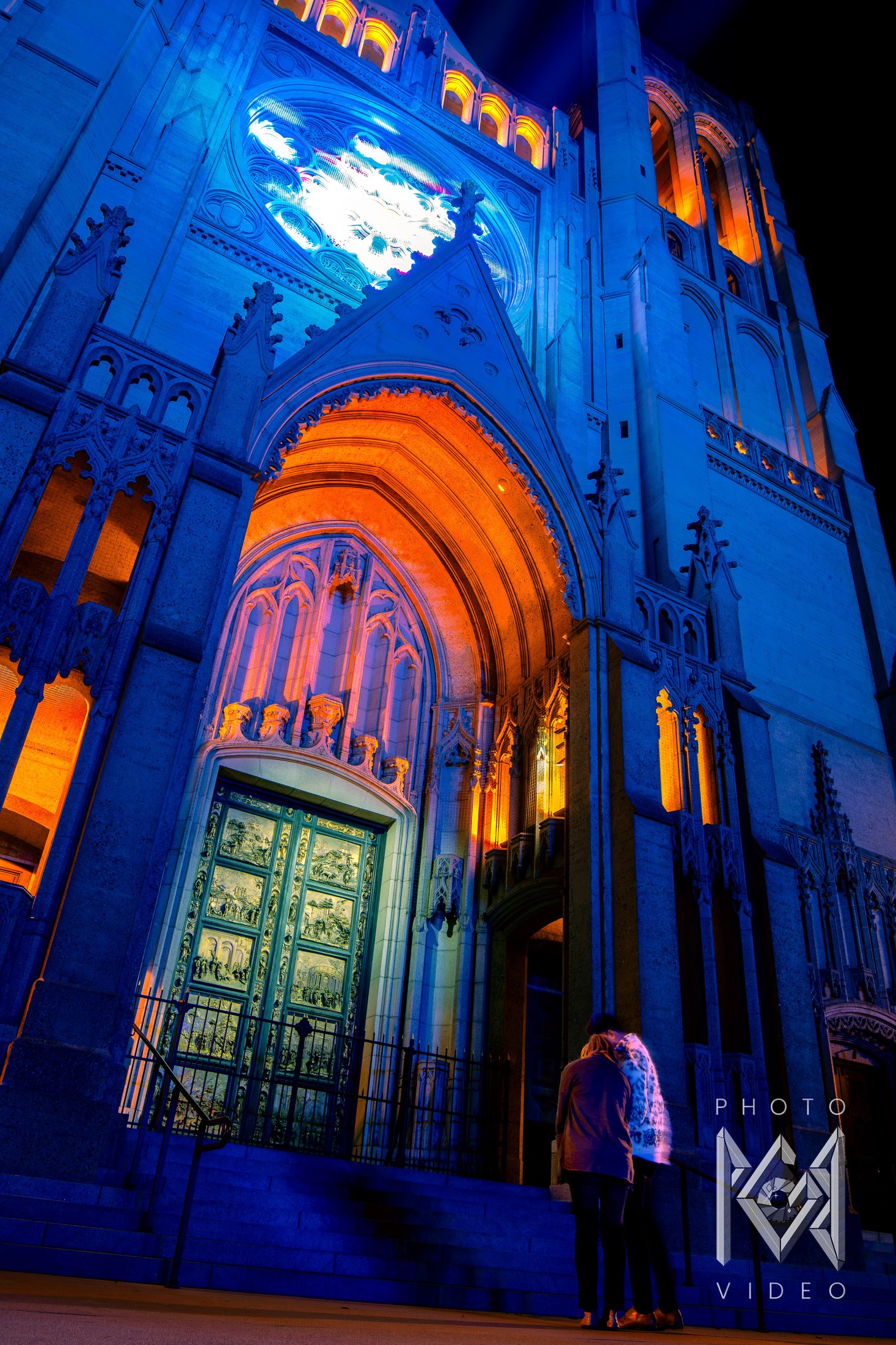 A couple embracing before a cathedral doorway, its carved arch glowing orange beneath a blue-lit facade and bright rose window