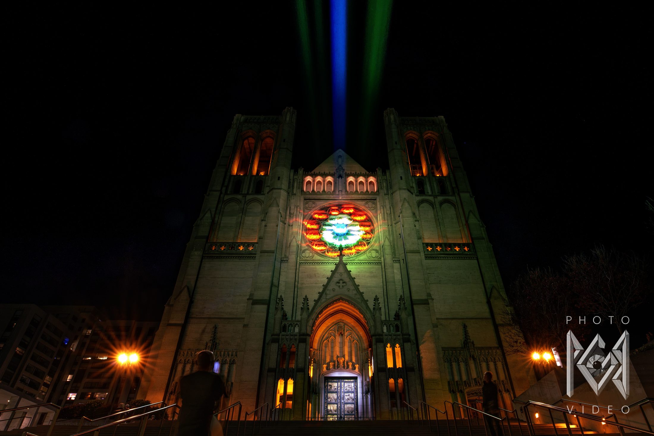 A cathedral front at night with a projected rose window, glowing bell towers and blue and green beams rising into the dark sky