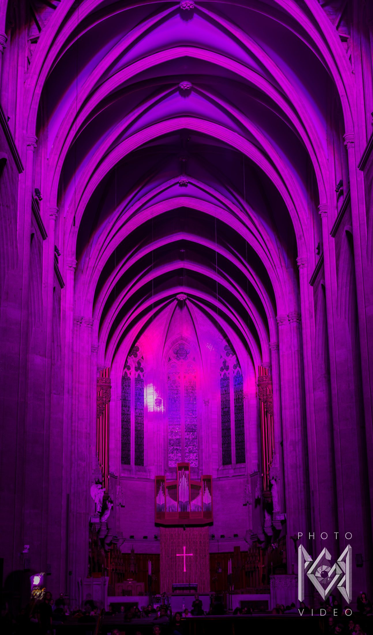 A cathedral nave washed in magenta and violet light, ribbed vaults leading to the glowing windows above the organ