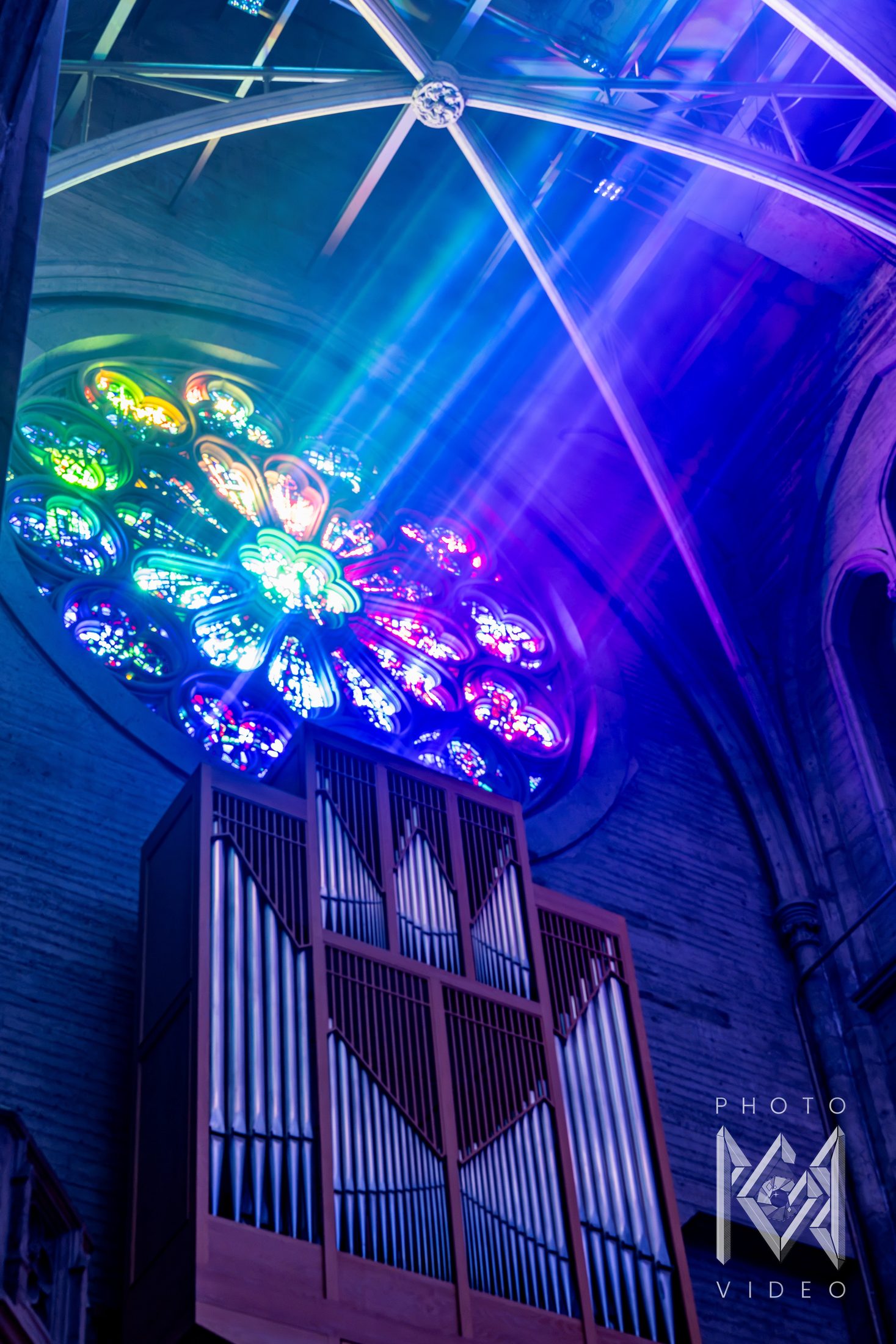 Beams of coloured light streaming from a rose window above the organ pipes, under ribs lit blue and violet