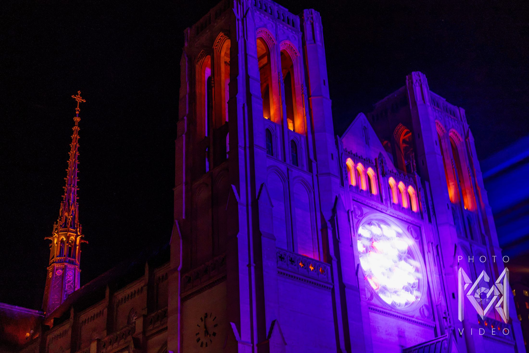 Cathedral bell towers and a gilded spire lit violet and orange against the night sky, beside a projected rose window