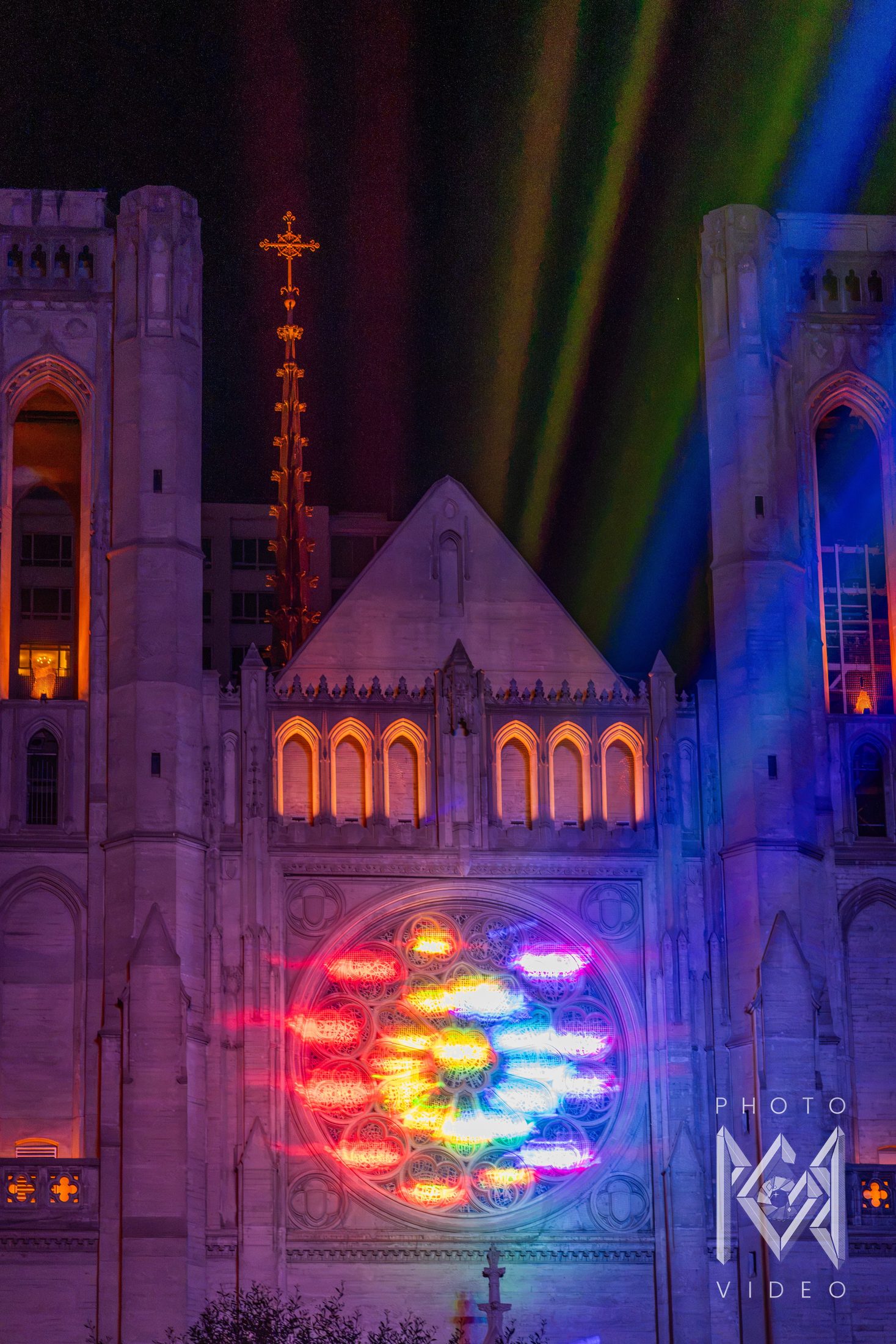 A cathedral facade with a rose window projected in rainbow light, beneath a gilded spire and bands of coloured light rising into the night