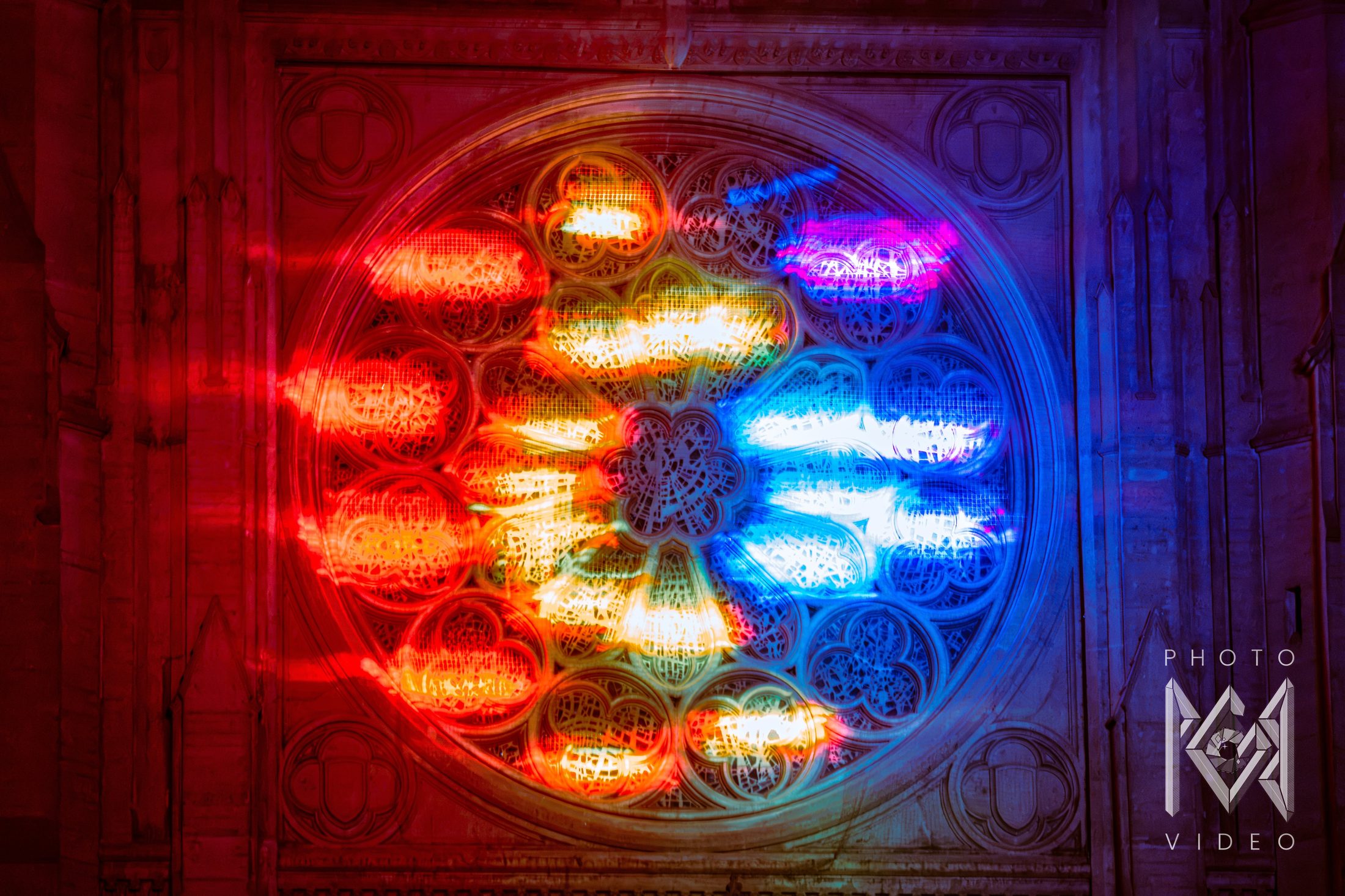 Close-up of a rose window lit in a sweep of red, gold, white and blue light across its stone tracery