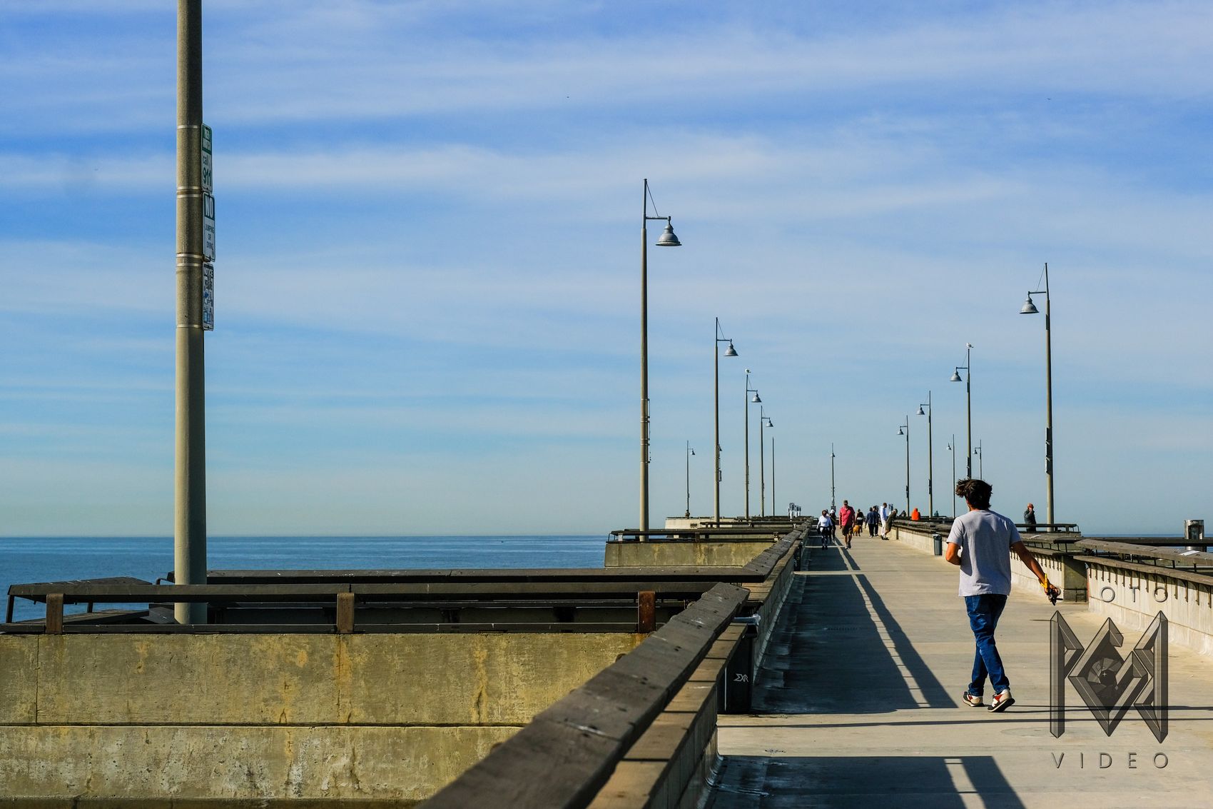 A young man in a grey T-shirt walking along a long pier lined with lampposts, people strolling ahead under a blue sky