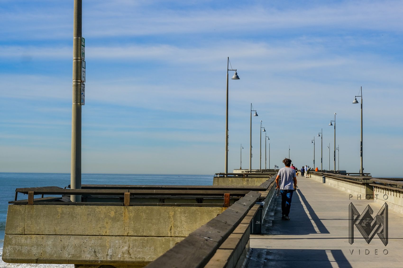 A young man walking away down the pier past its tall lampposts, the ocean stretching out to the left