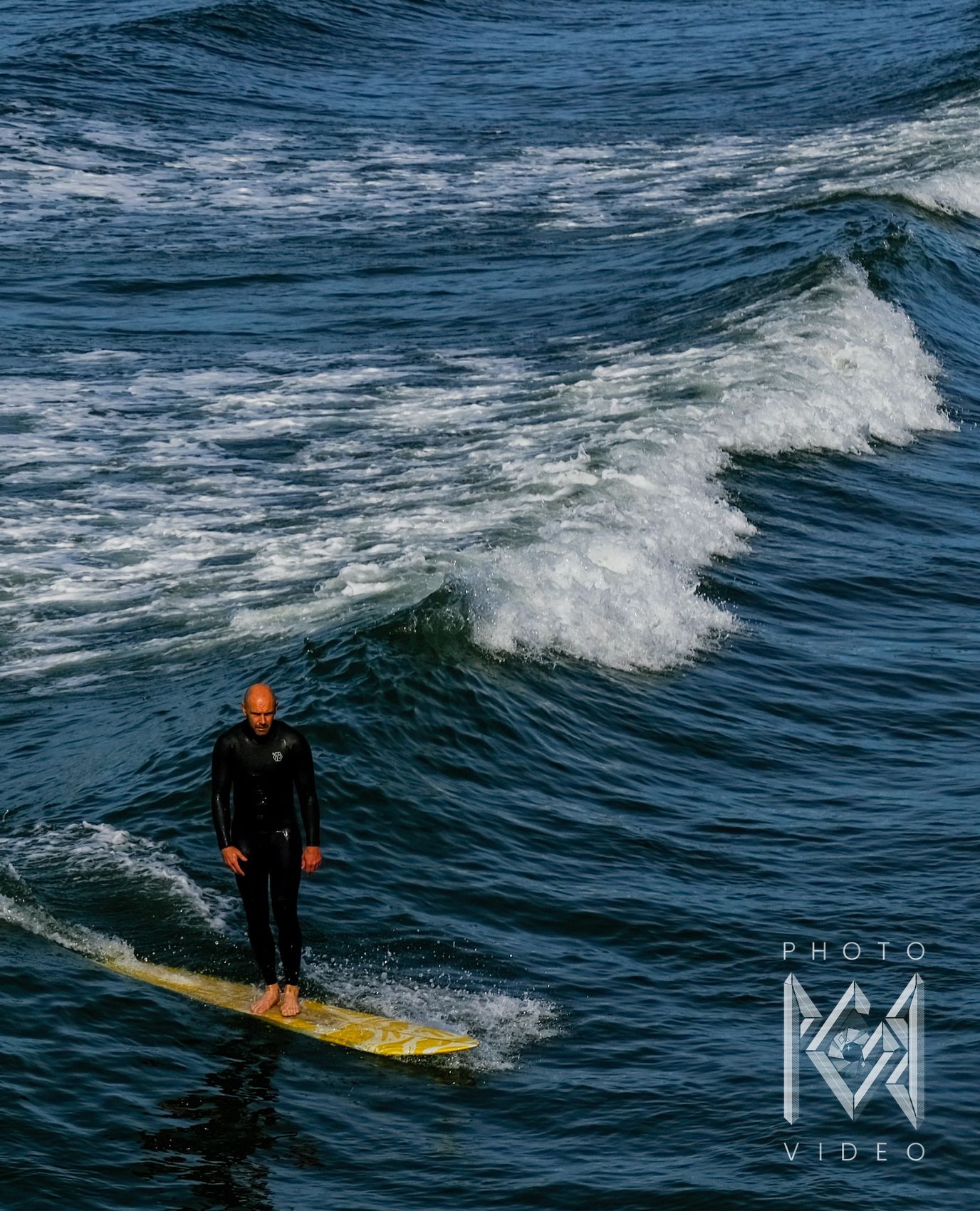 A bald surfer in a black wetsuit riding a yellow longboard across deep blue water, a wave breaking behind him