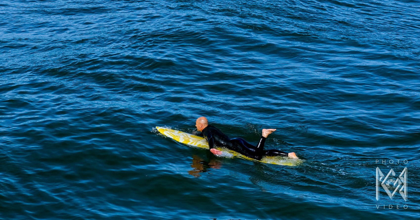 A surfer in a black wetsuit lying on a yellow board, paddling across rippled blue water