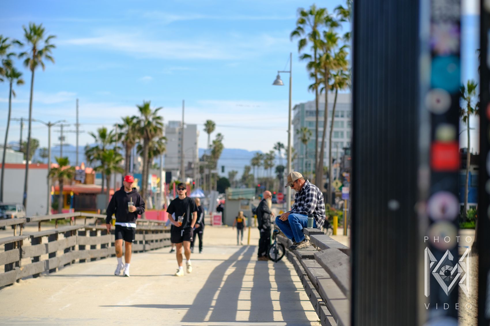People walking and sitting along a sunny boardwalk lined with palm trees, shops and lampposts