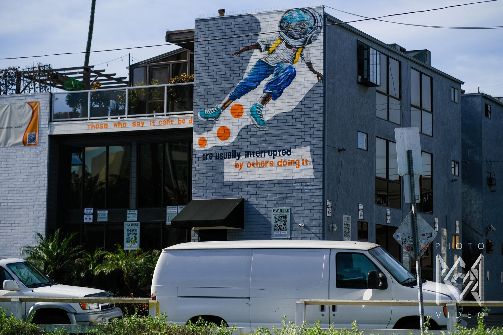 A mural of a figure in an astronaut helmet leaping across a brick building, above the words "are usually interrupted by others doing it"