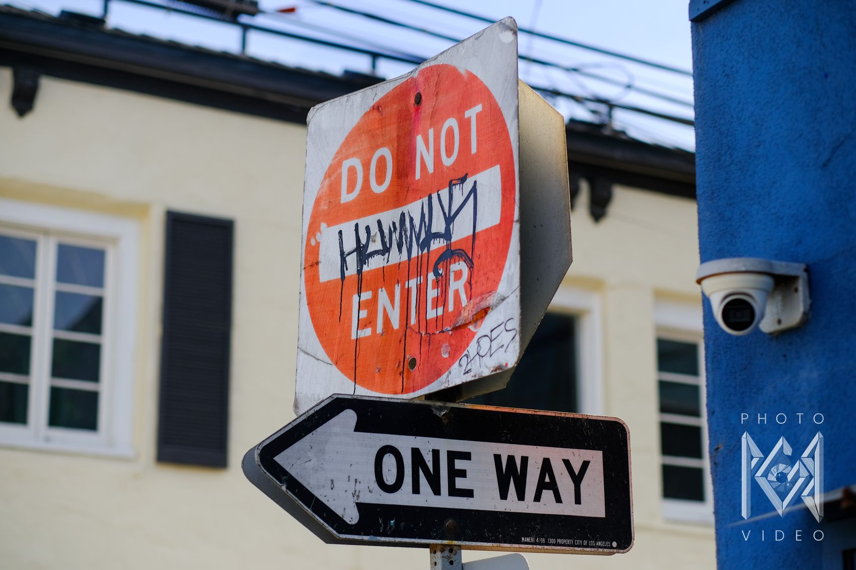 A weathered Do Not Enter sign tagged with graffiti above a One Way arrow, with a security camera on a blue wall beside it