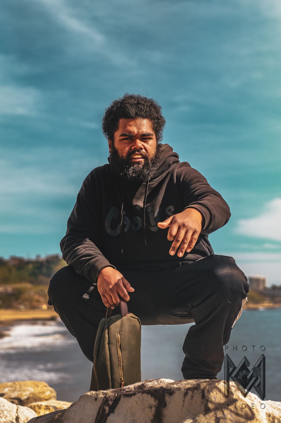 A bearded man in a black hoodie crouching on a rock by the sea, one hand gesturing toward the camera, under a teal sky