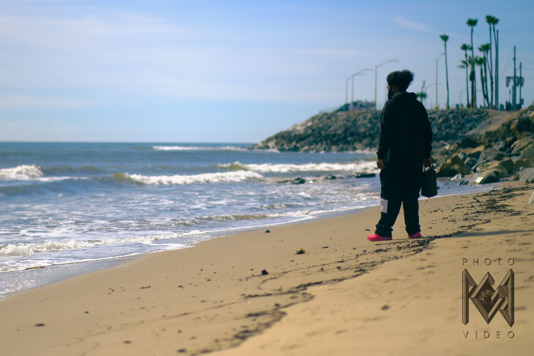 A man in a black hoodie and pink shoes standing at the water's edge, looking out along the beach toward a rocky point and palm trees