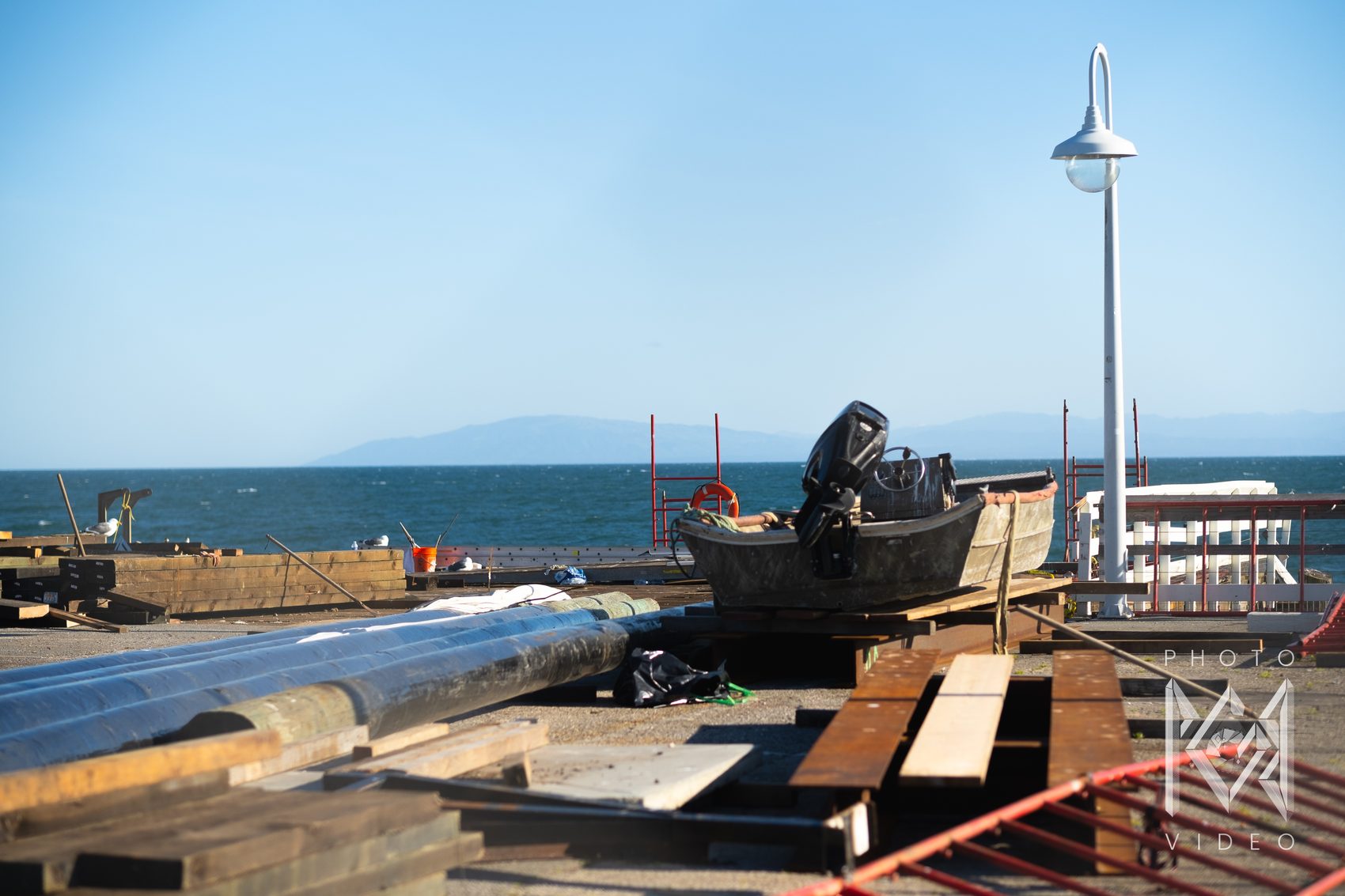A small boat and its outboard motor set up on a pier among stacked timbers and pilings under repair, the bay and distant mountains beyond