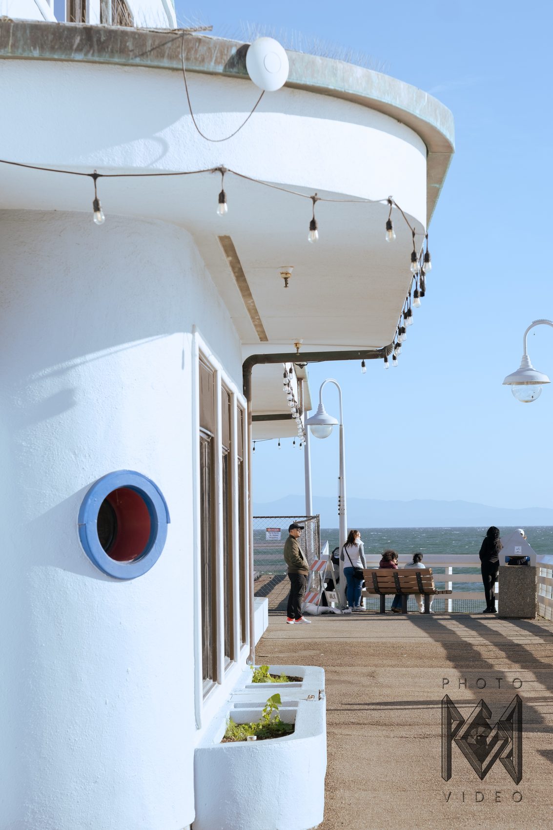 A curved white pier building with a blue porthole window and string lights, people resting at the railing beyond
