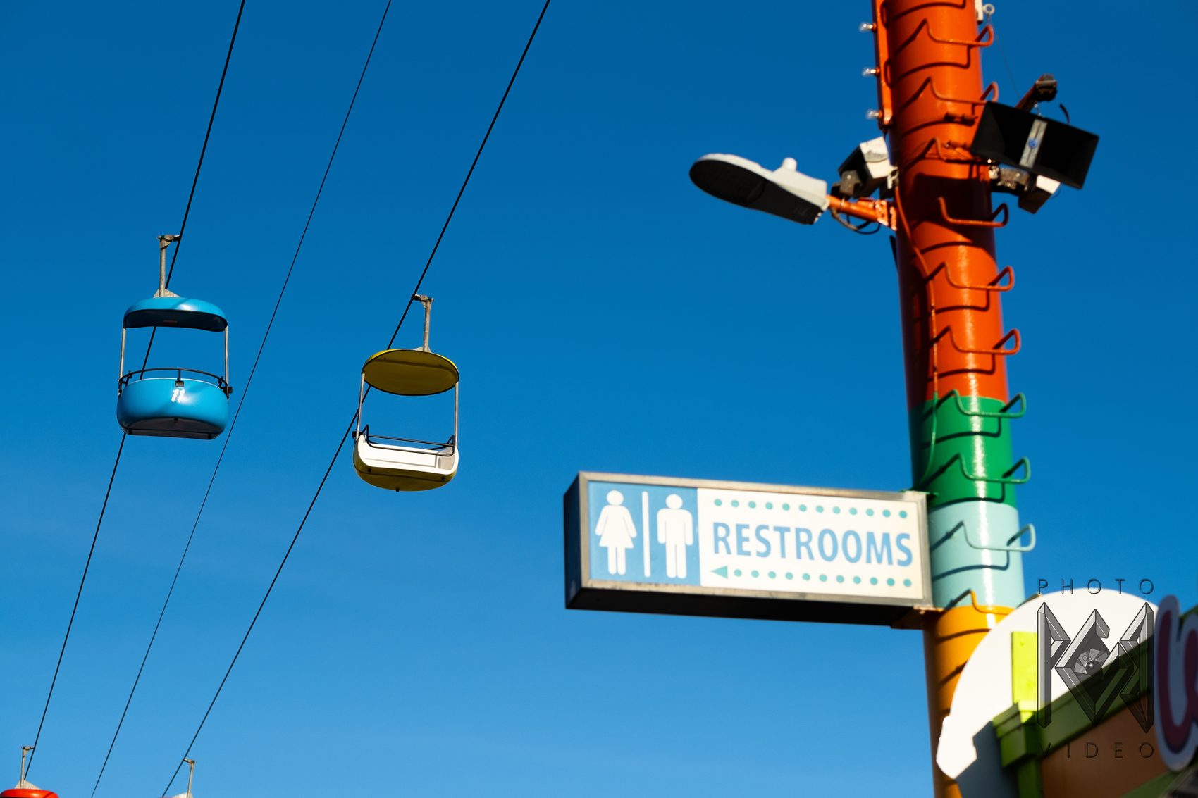 Blue and yellow sky ride gondolas on their cables beside an orange striped pole and a Restrooms sign, against a clear blue sky