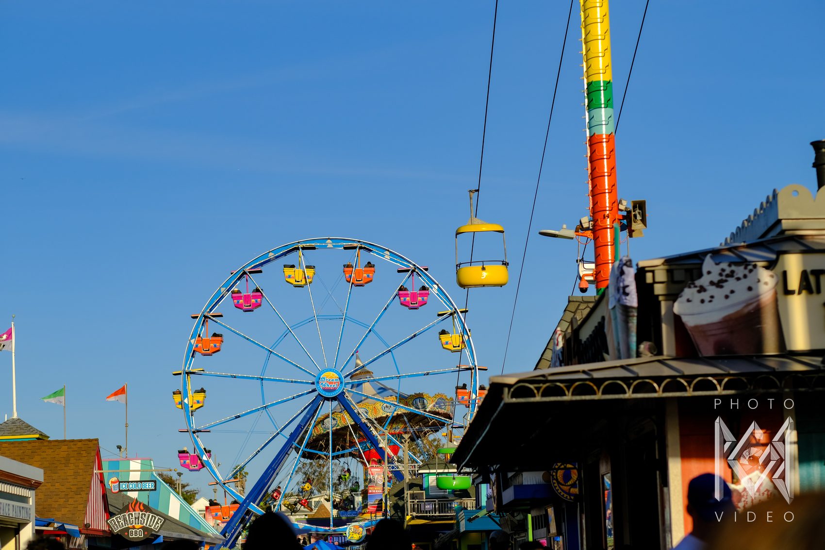 A blue Ferris wheel with bright pink, yellow and orange cars rising over the boardwalk shops, a yellow sky ride gondola passing by