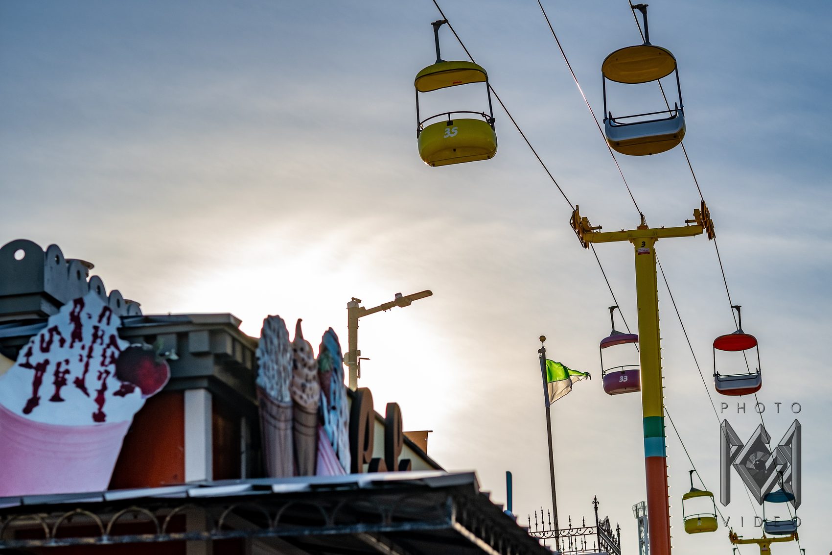 Sky ride gondolas in yellow, pink and red gliding past an ice-cream stand in soft late light