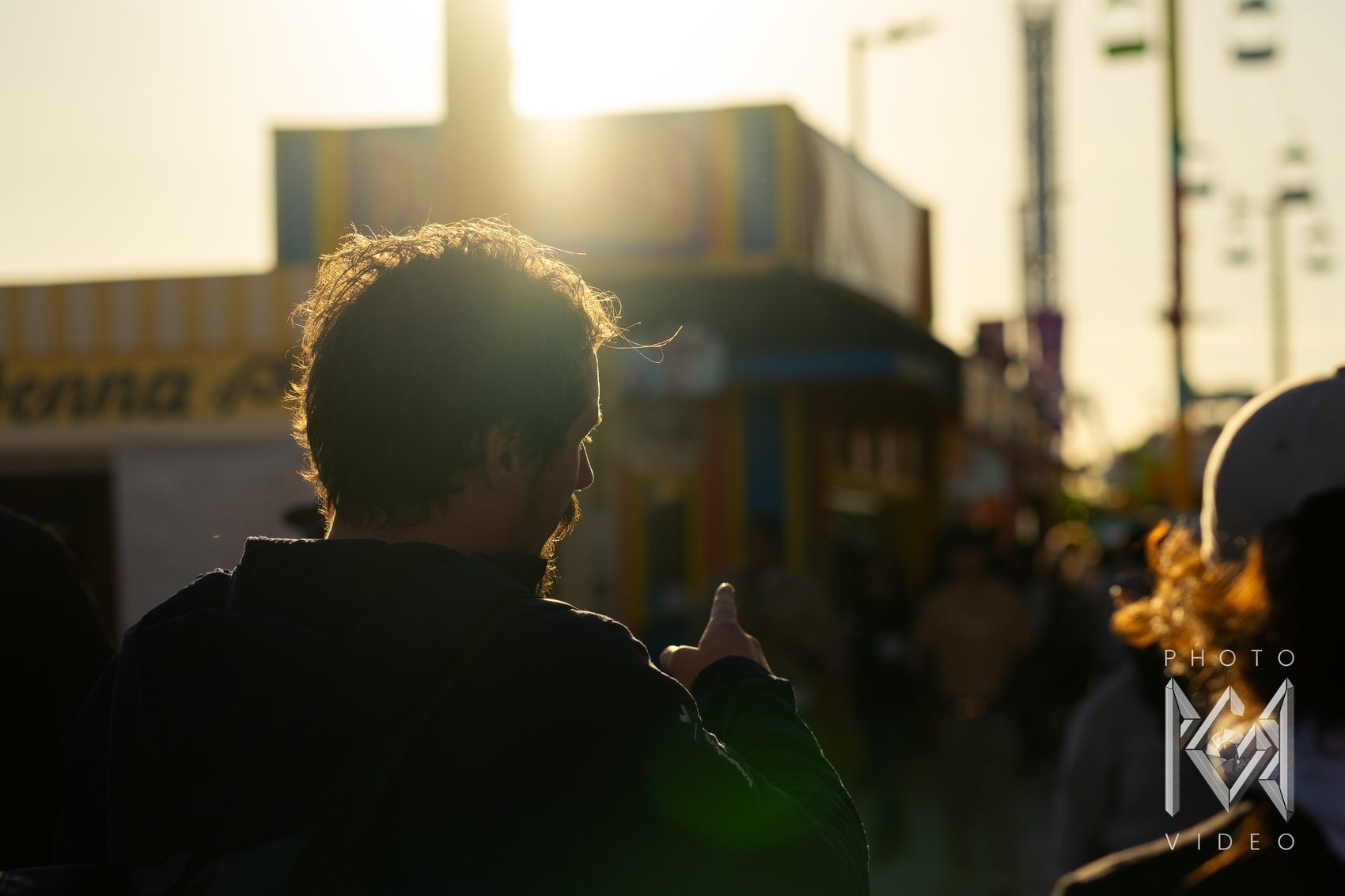 A young man seen from behind, his hair lit gold by the low sun, pointing ahead along the busy boardwalk