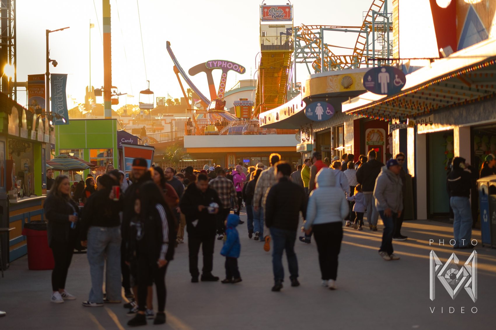 Crowds strolling the boardwalk at sunset beneath the Typhoon ride and a roller coaster, the shops glowing gold
