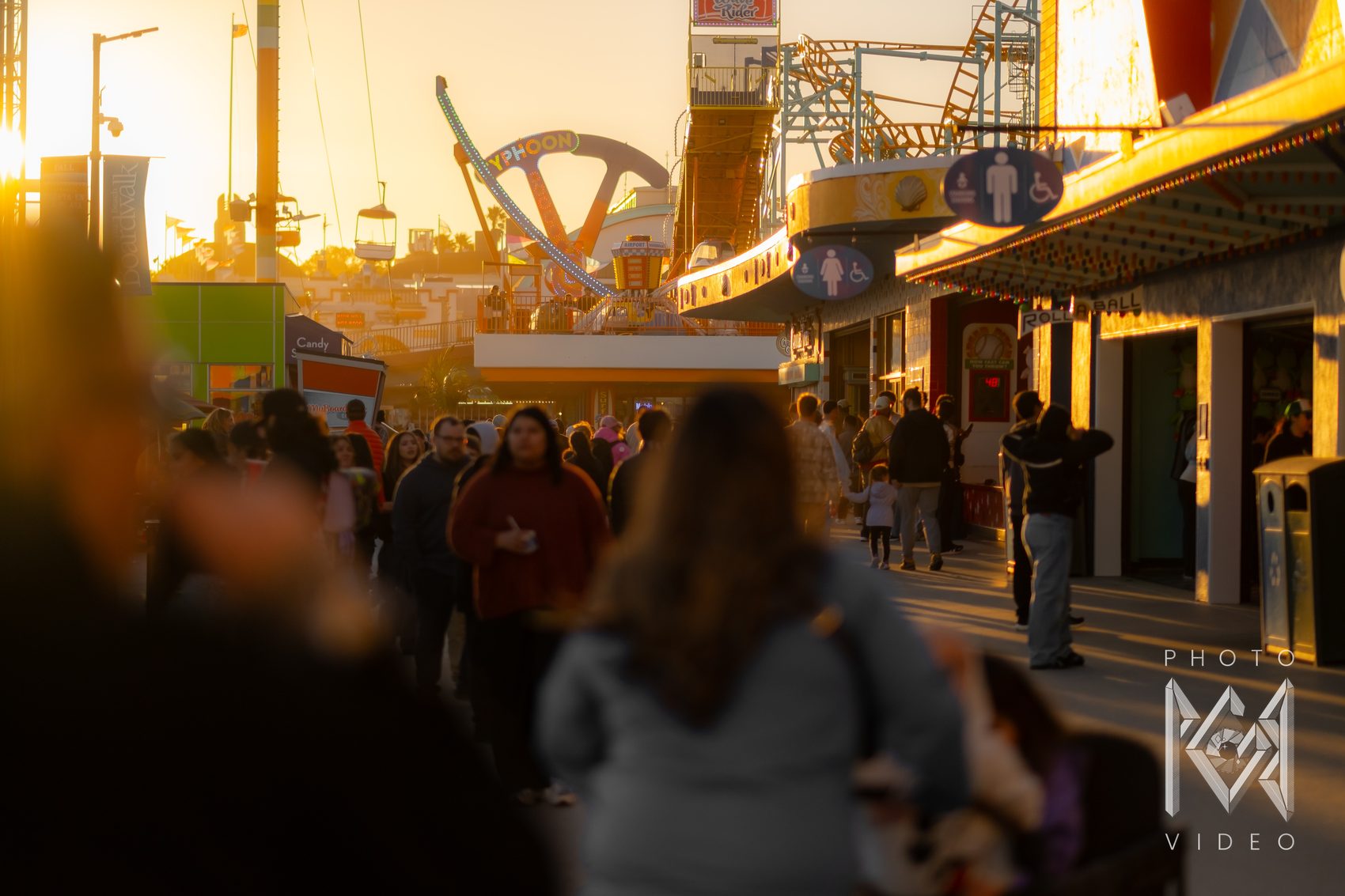 The same boardwalk crowd at golden hour, soft blurred figures in the foreground and the Typhoon ride beyond