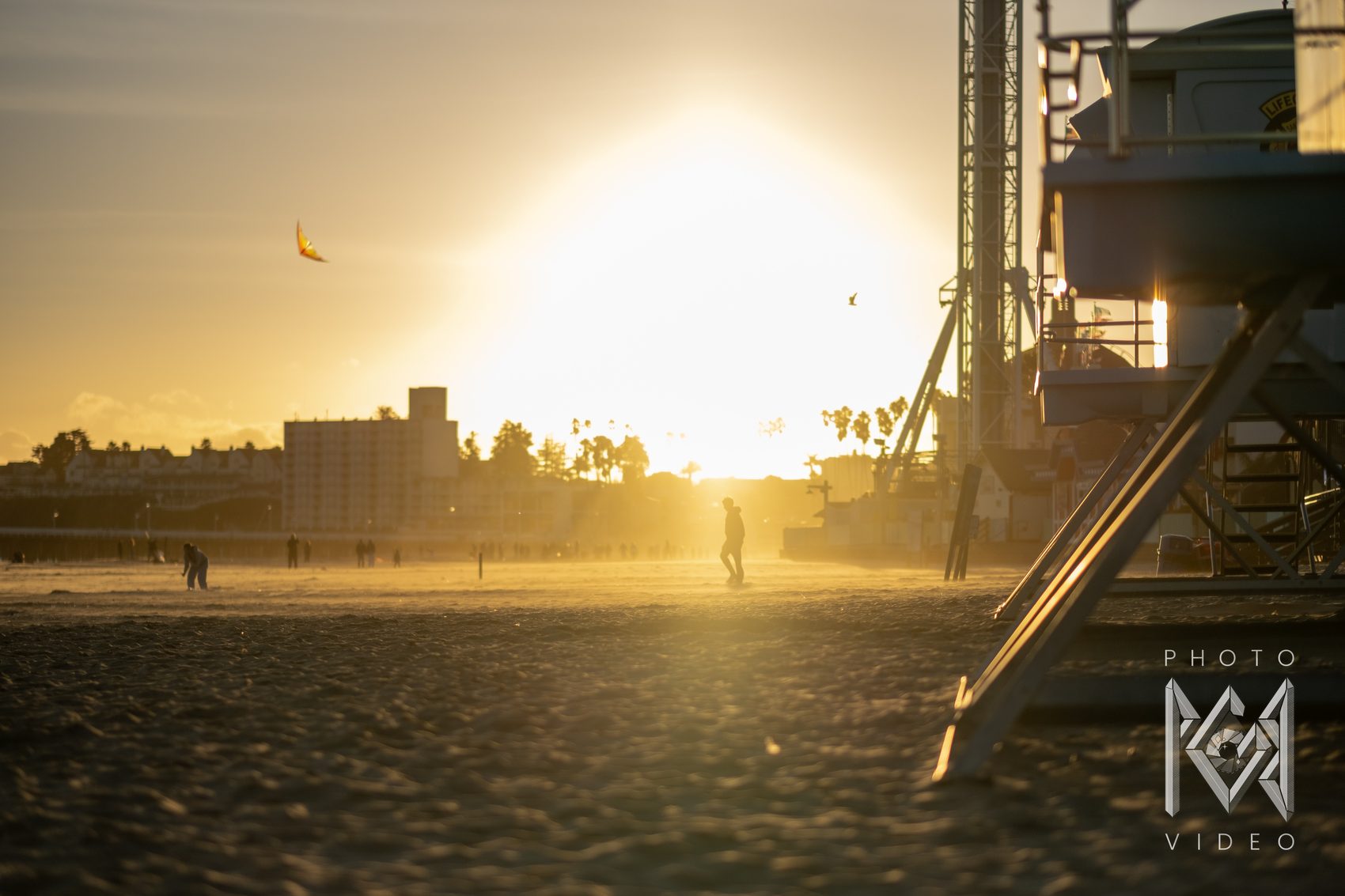 A lone walker crossing the sand in a golden haze at sunset, a kite overhead and a lifeguard tower to the side