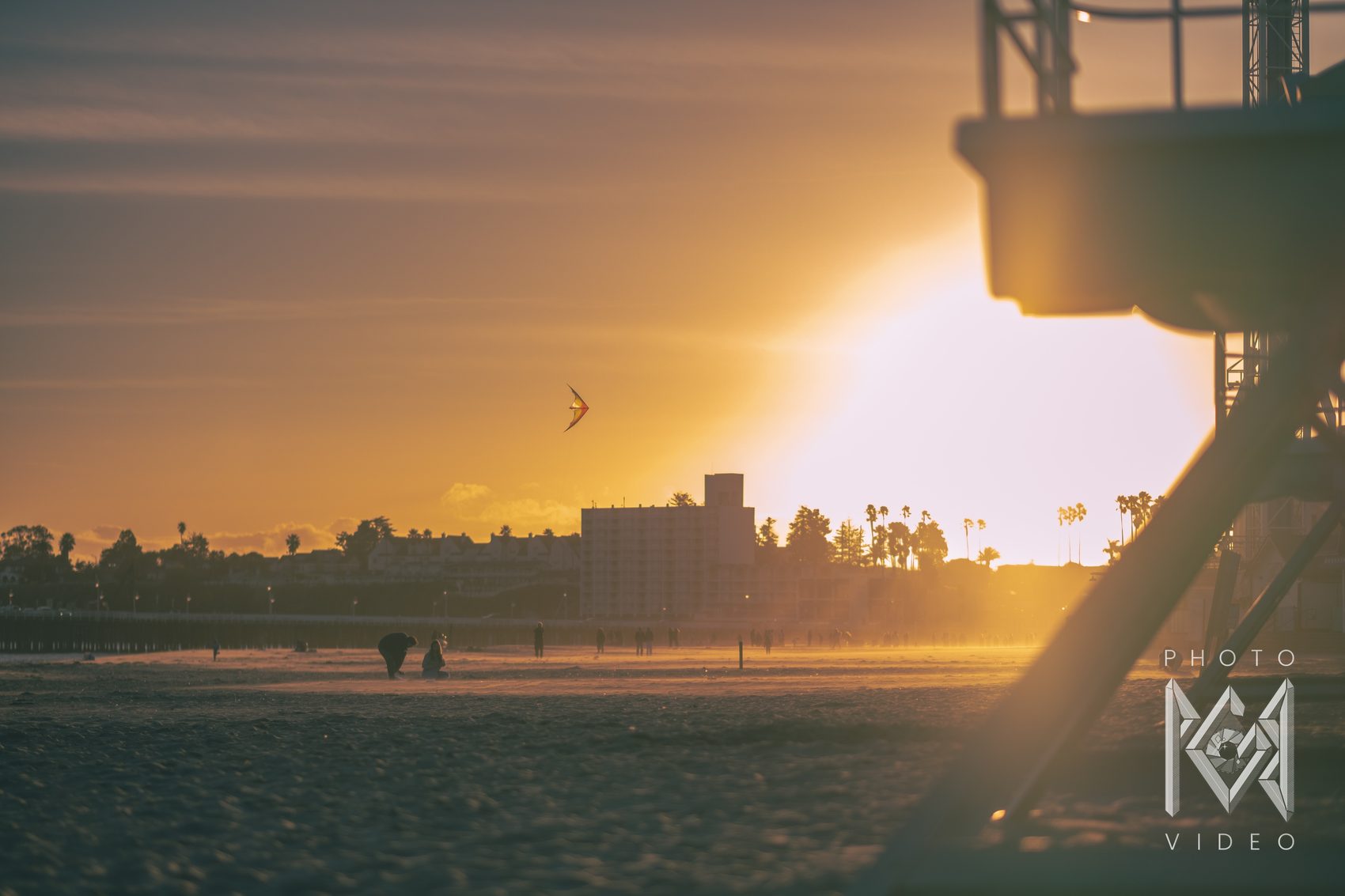 A stunt kite against an amber sunset sky over the beach, people silhouetted on the sand beside a lifeguard tower