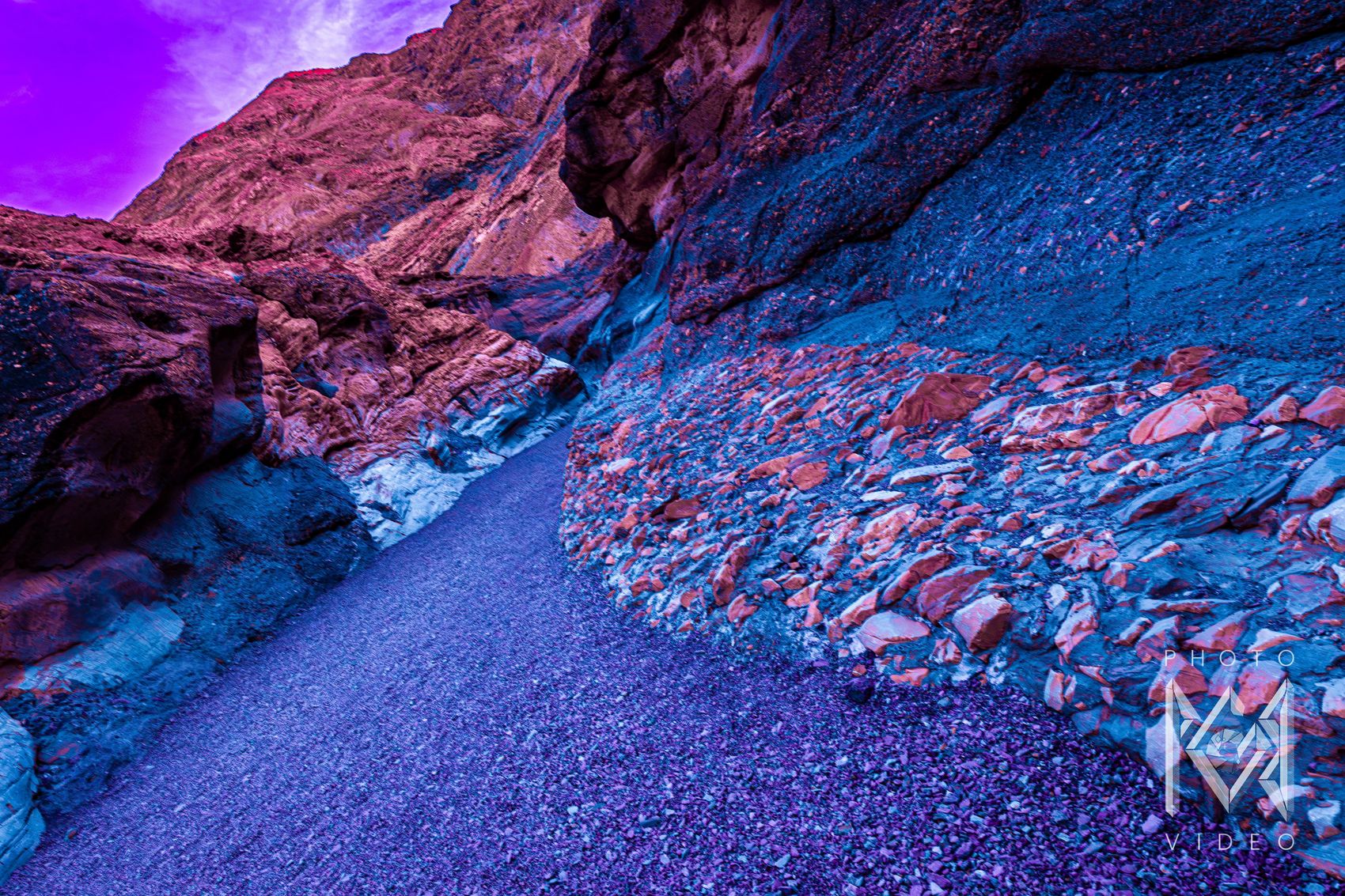 A winding canyon path of violet gravel between rose-red cliffs and a blue slope strewn with coral-pink stones, under a magenta sky