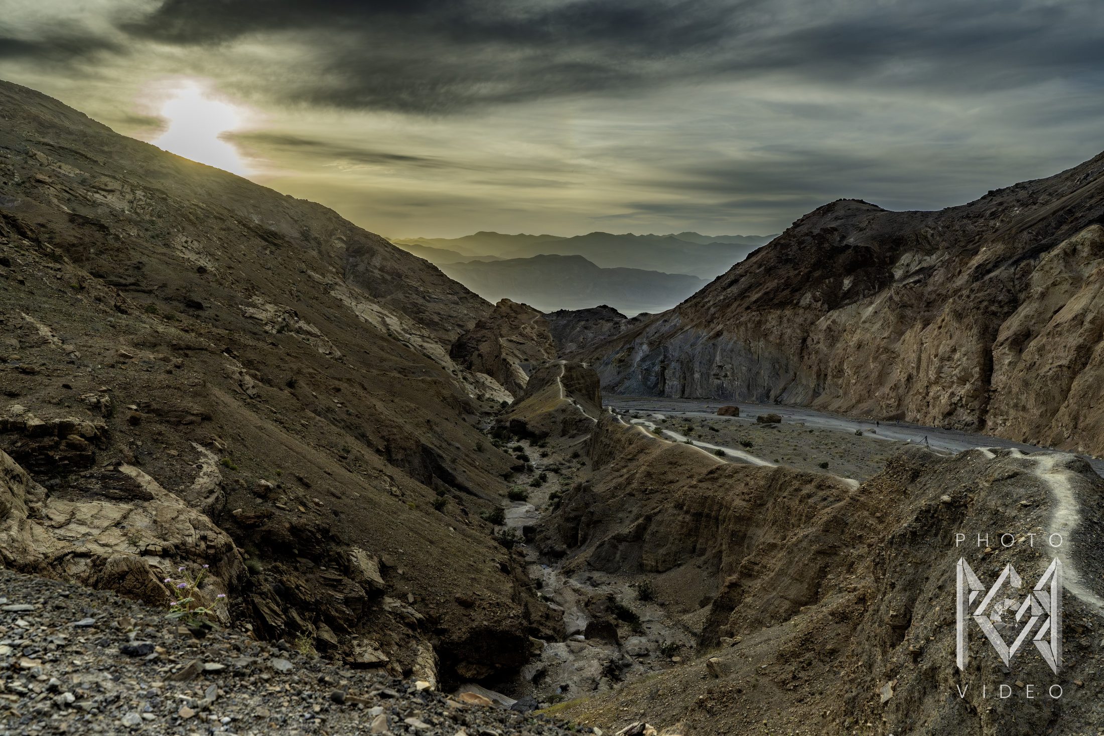 A winding desert canyon with a footpath along its rim, rocky slopes on both sides, hazy mountain ranges and a low sun behind dark clouds