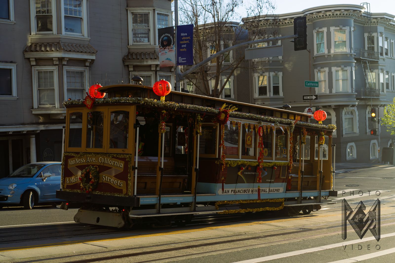 A wooden cable car trimmed with red lanterns climbing a sunlit street past bay-windowed Victorian houses
