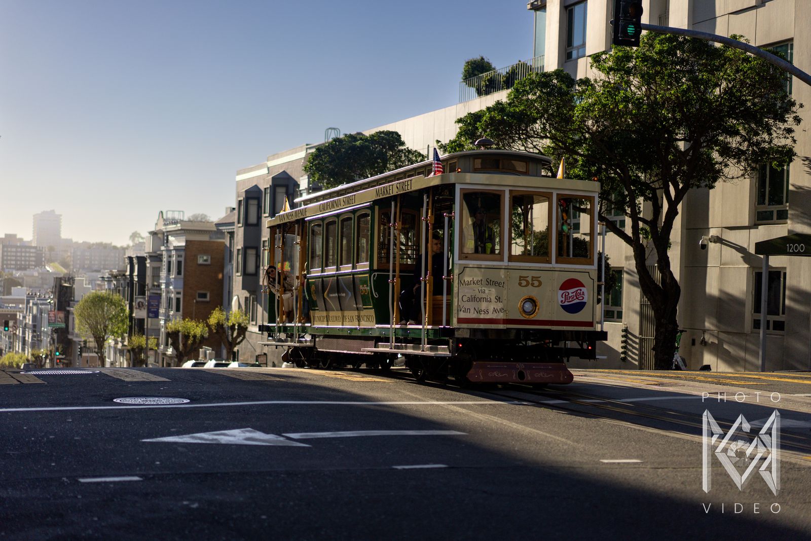 Cable car 55 cresting a steep hill in afternoon light, trees and rows of houses falling away to the bay behind it