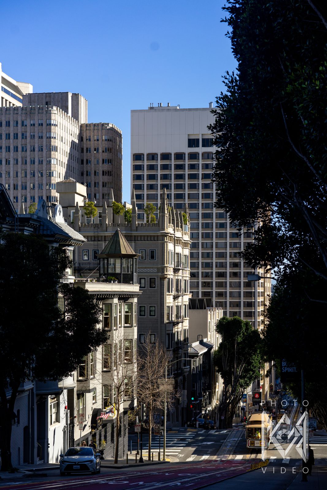 A steep street dropping away between tall buildings and dark trees, a small cable car far below in the sun