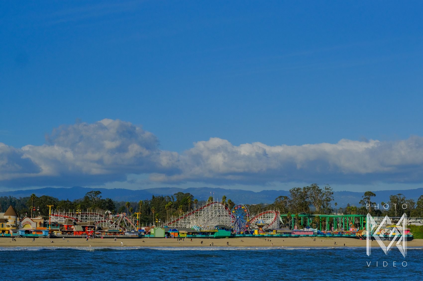 A white wooden roller coaster, a blue Ferris wheel and colourful rides along the beach, seen from the water