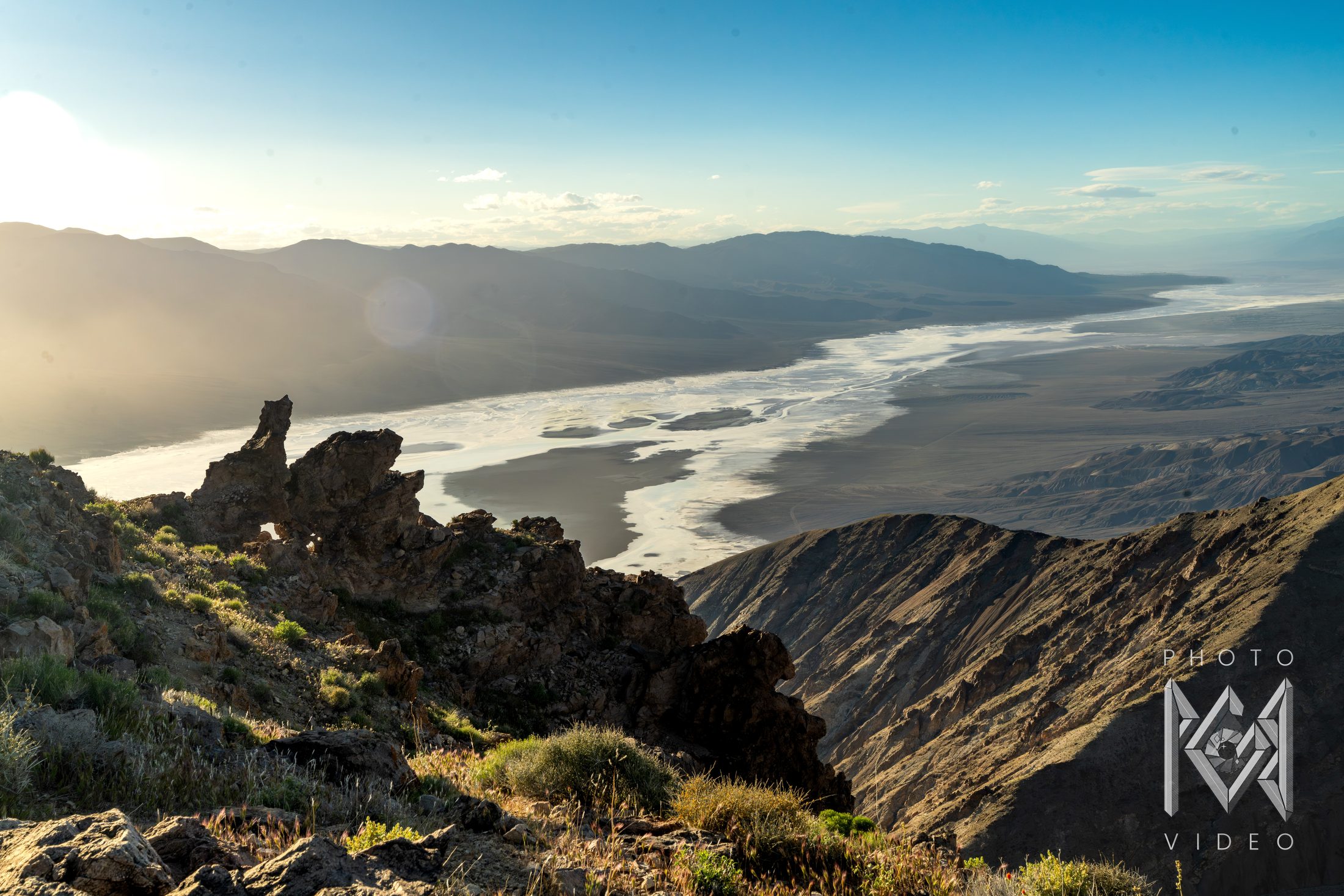 Late sun over a desert valley, its white salt flats winding between mountain ranges, seen past a jagged rocky ridge